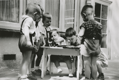 876986 Afbeelding van een groepje kleuters dat kijkt naar de kleinste kleuter knutselend aan een tafeltje op de ...